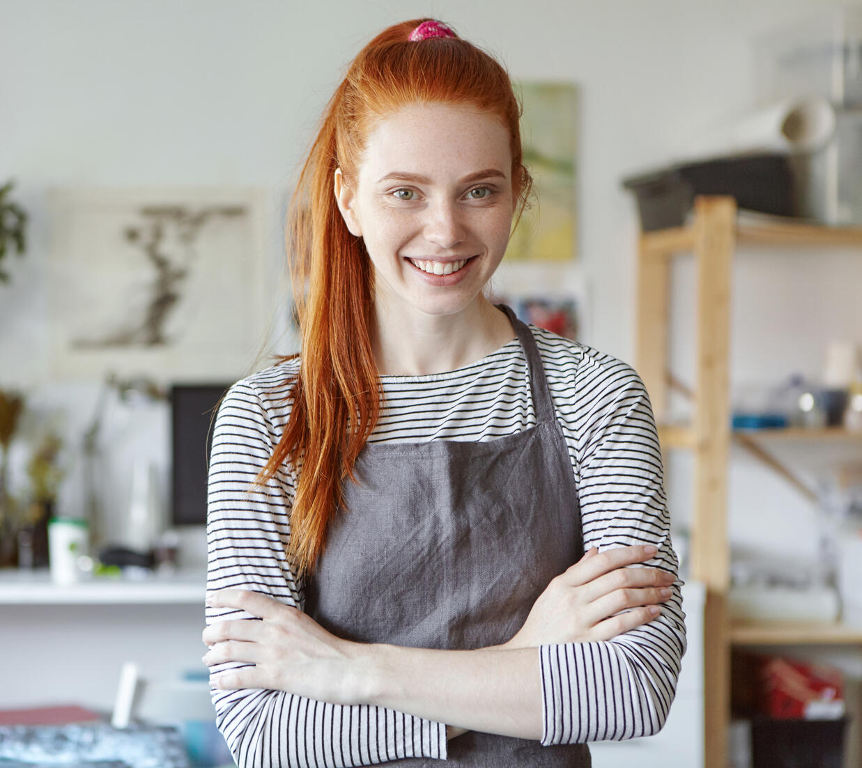 Smiling young small business owner wearing an apron, standing confidently in a bright creative studio filled with plants and art supplies.