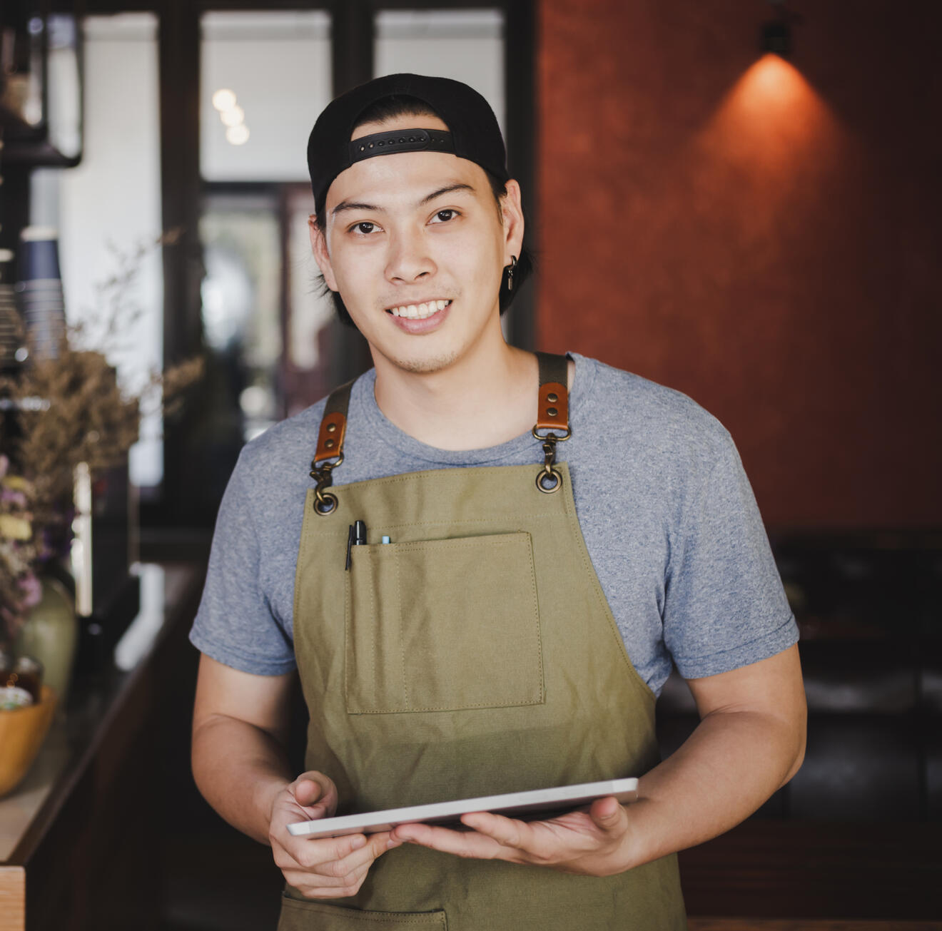 Smiling small-business owner wearing a green apron and holding a tablet in a cozy café or workshop filled with handmade goods and plants.