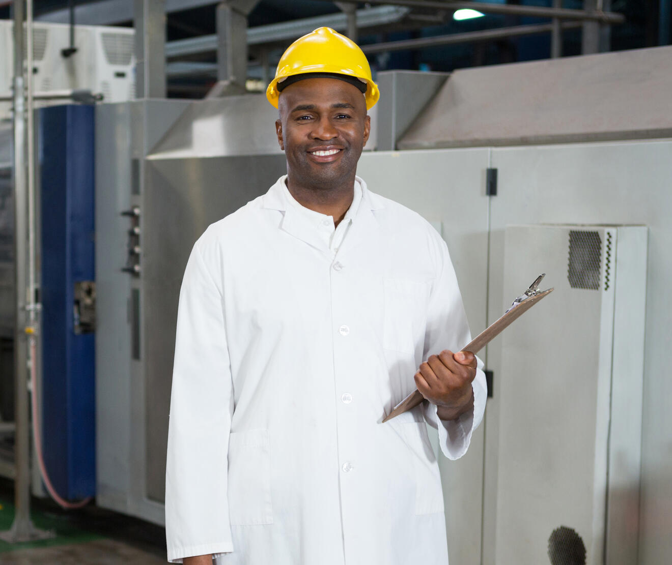 Factory worker wearing a white lab coat and yellow hard hat, standing in front of industrial machines and holding a clipboard with a confident smile.